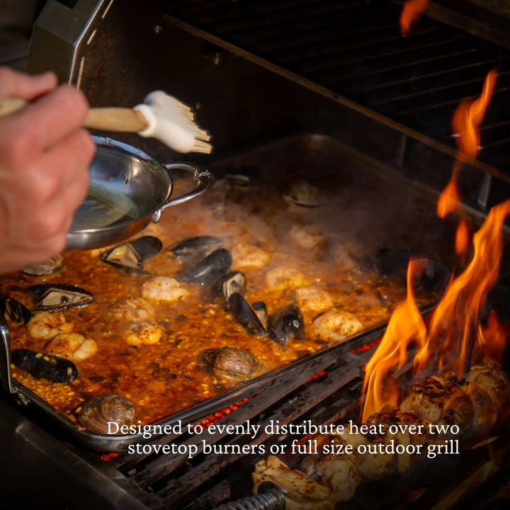 A person brushes sauce onto seafood paella cooking on a Hestan Provisions Double Burner Griddle (21"x13") over an outdoor grill. Text reads: Designed to evenly distribute heat over two stovetop burners or a double burner griddle.