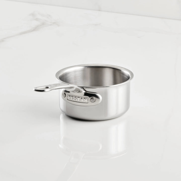A small stainless steel saucepan from the Hestan Provisions Holiday Host and Roast 8-piece Set, featuring a short handle engraved with "NESTAN," sits clean and empty on a white marble surface as part of a durable bakeware collection.