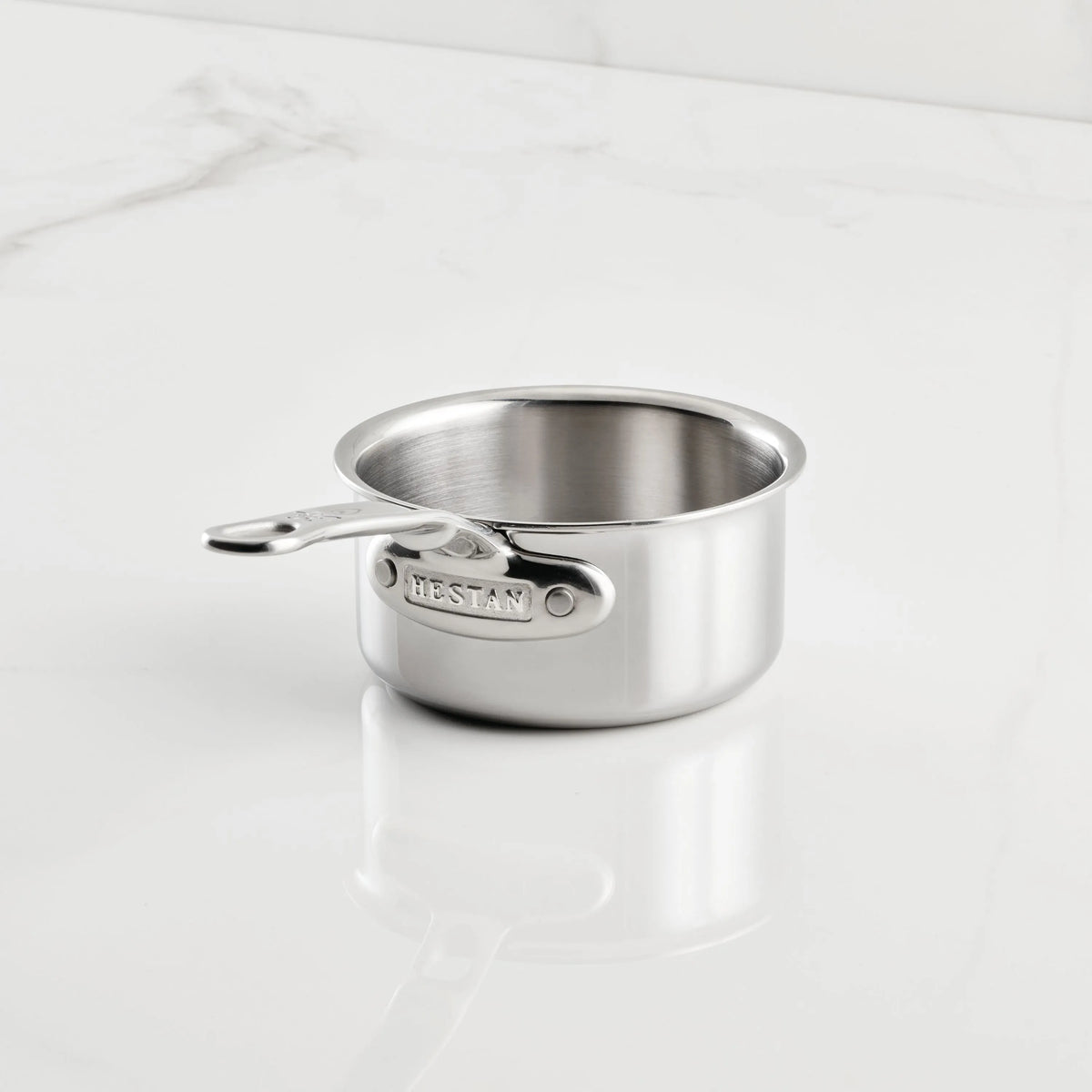 A small stainless steel saucepan from the Hestan Provisions Host and Roast 8-piece set rests empty on a white marble surface, with the brand name engraved on its short handle.
