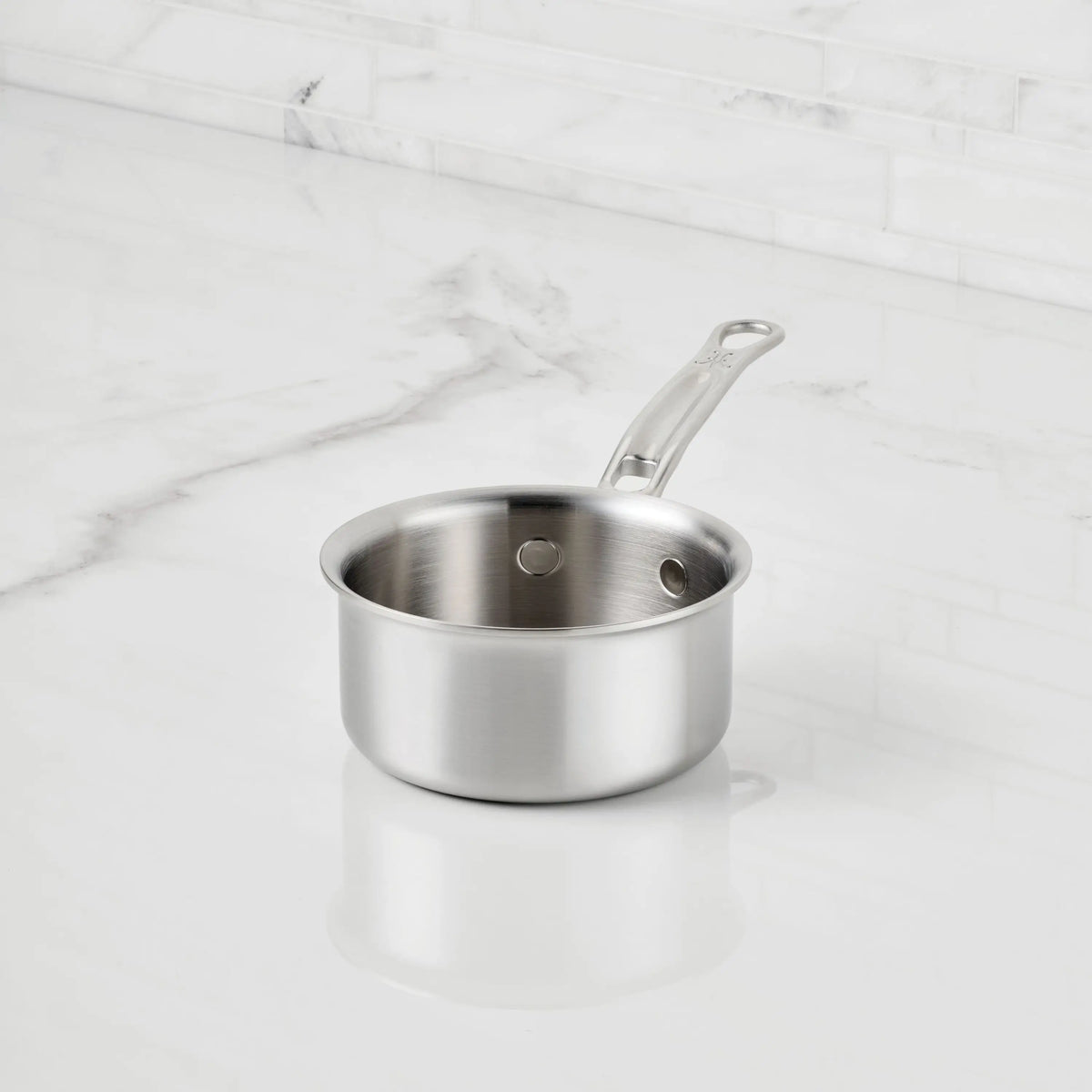 The ProBond® Luxe™ Polished Clad Stainless Steel Butter Warmer, 0.75-Quart, with a long handle, sits on a white marble countertop, highlighting its Italian craftsmanship against a tiled backsplash.