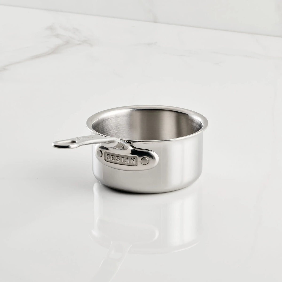 A compact ProBond® Luxe™ Polished Clad Stainless Steel Butter Warmer, 0.75-quart, with a short handle sits on white marble, highlighting its exquisite Italian craftsmanship.