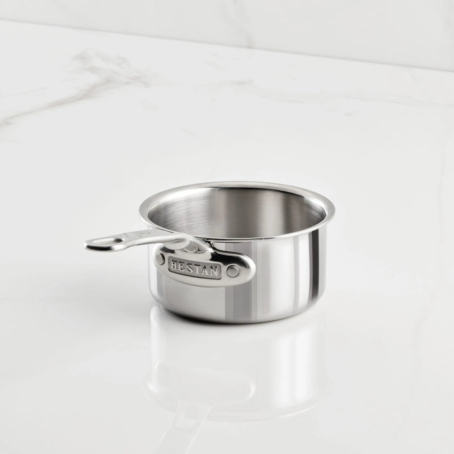 A ProBond Luxe® Polished Clad Stainless Steel Butter Warmer, 0.75-quart, rests on a white marble surface with subtle gray veining. The butter warmer features a polished, reflective finish and is empty.