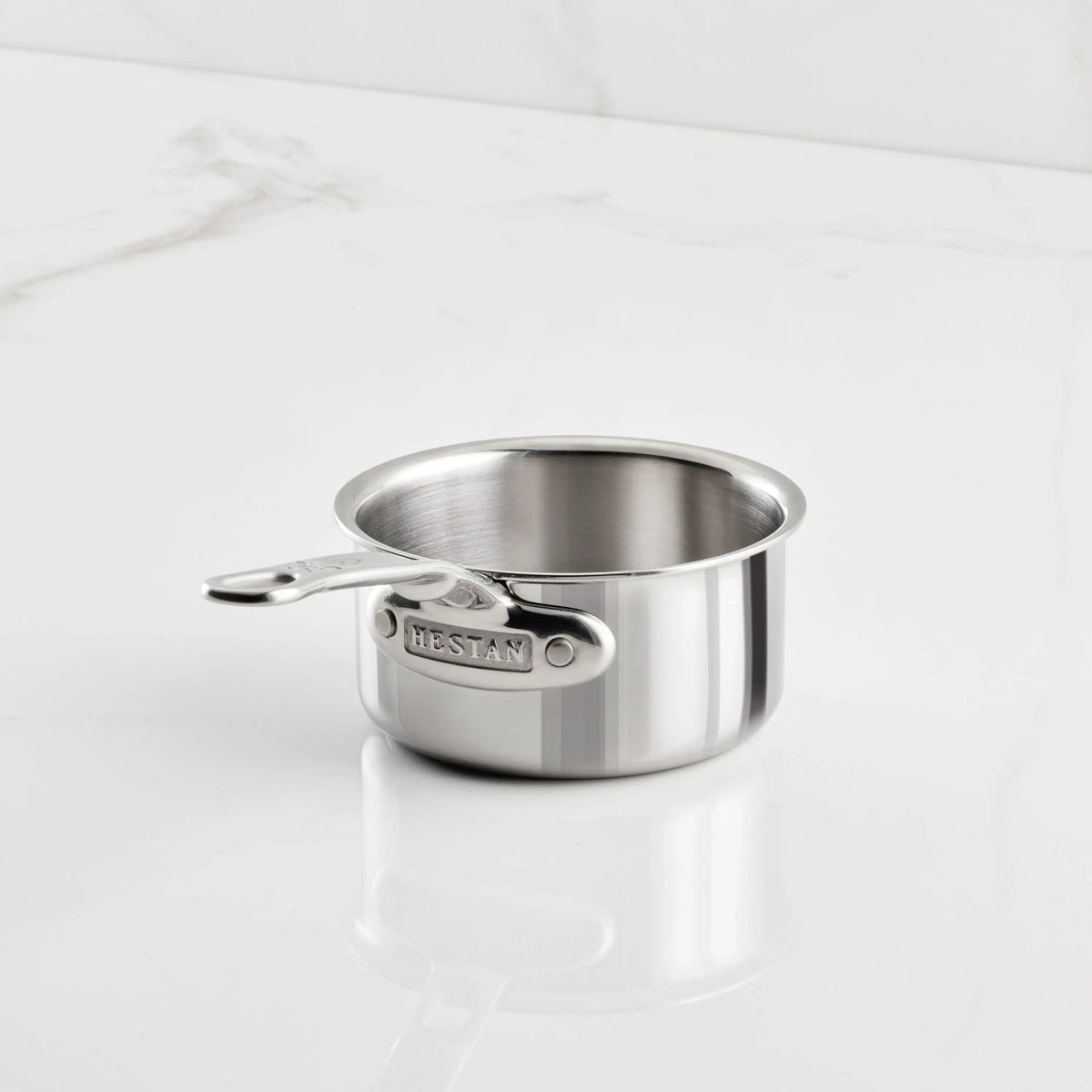 A ProBond Luxe® Polished Clad Stainless Steel Butter Warmer, 0.75-quart, rests on a white marble surface with subtle gray veining. The butter warmer features a polished, reflective finish and is empty.