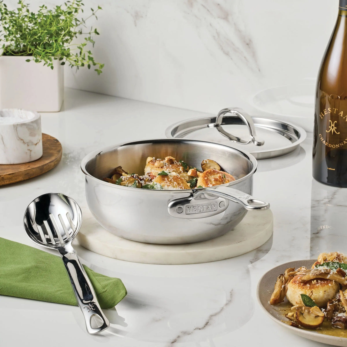 The ProBond® Luxe™ Polished Clad Stainless Steel Signature Set, 7-Piece, sits on a marble trivet with matching lid and serving spoon nearby, showcasing its induction-ready versatility on a white countertop.