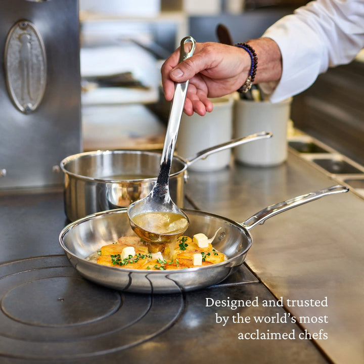 A chef pours broth into a ProBond® Luxe™ Polished Clad Stainless Steel Soup Pot with Lid filled with vegetables and herbs on the stove. A saucepan and utensils are nearby. Text: “Designed and trusted by the world’s most acclaimed chefs.”.