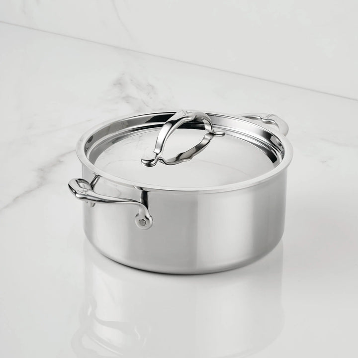 The ProBond® Luxe™ Polished Clad Stainless Steel Soup Pot with Lid features Italian craftsmanship, two handles, and a large curved lid handle. It sits on a white marble surface, gleaming with its shiny finish.
