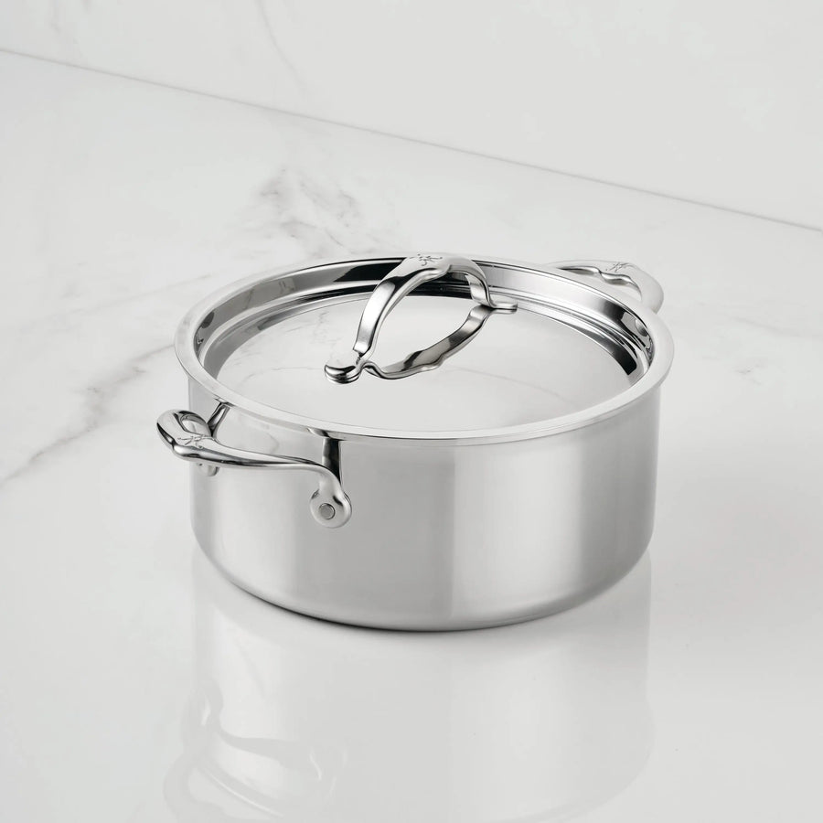The ProBond® Luxe™ Polished Clad Stainless Steel Soup Pot with Lid features Italian craftsmanship, two handles, and a large curved lid handle. It sits on a white marble surface, gleaming with its shiny finish.
