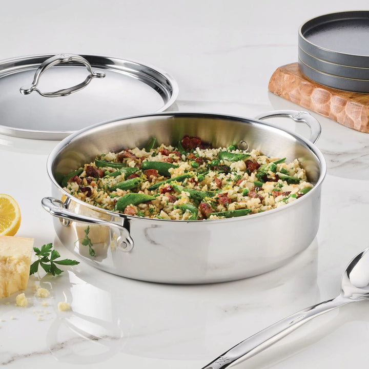 A ProBond® Luxe™ Polished Clad Stainless Steel Rondeau filled with mixed vegetables and grains rests on a marble countertop, with its matching lid, serving spoon, gray plates, cheese, herbs, and a lemon wedge nearby.