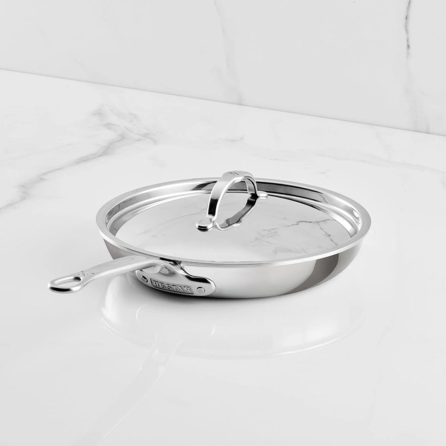 A ProBond Luxe® Polished Clad Stainless Steel Covered Skillet rests on a white marble countertop. The skillet features a long handle and a metal loop atop its lid, reflecting the durability and elegance of tri-ply clad cookware. A plain white background with subtle marble veining completes the scene.