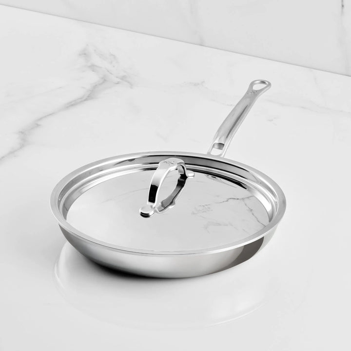 A Polished Clad Stainless Steel Covered Skillet from ProBond Luxe® rests on a white marble countertop. This cookware piece features a long handle, while the matching lid includes a sturdy loop handle. Both the skillet and lid boast a polished, reflective finish that showcases professional-quality craftsmanship.