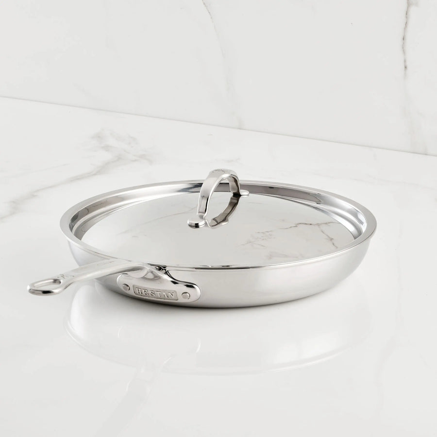 A ProBond® Luxe™ Polished Clad Stainless Steel Covered Skillet, with lid and handle, sits on a white marble countertop, highlighting the modern elegance of tri-ply cookware in contemporary kitchens.