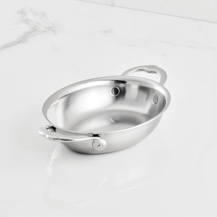 An 8.5-inch x 6.5-inch Hestan Provisions Oval Au Gratin pan with two riveted handles rests on a white marble surface, showcasing its shiny stainless steel finish and empty interior.