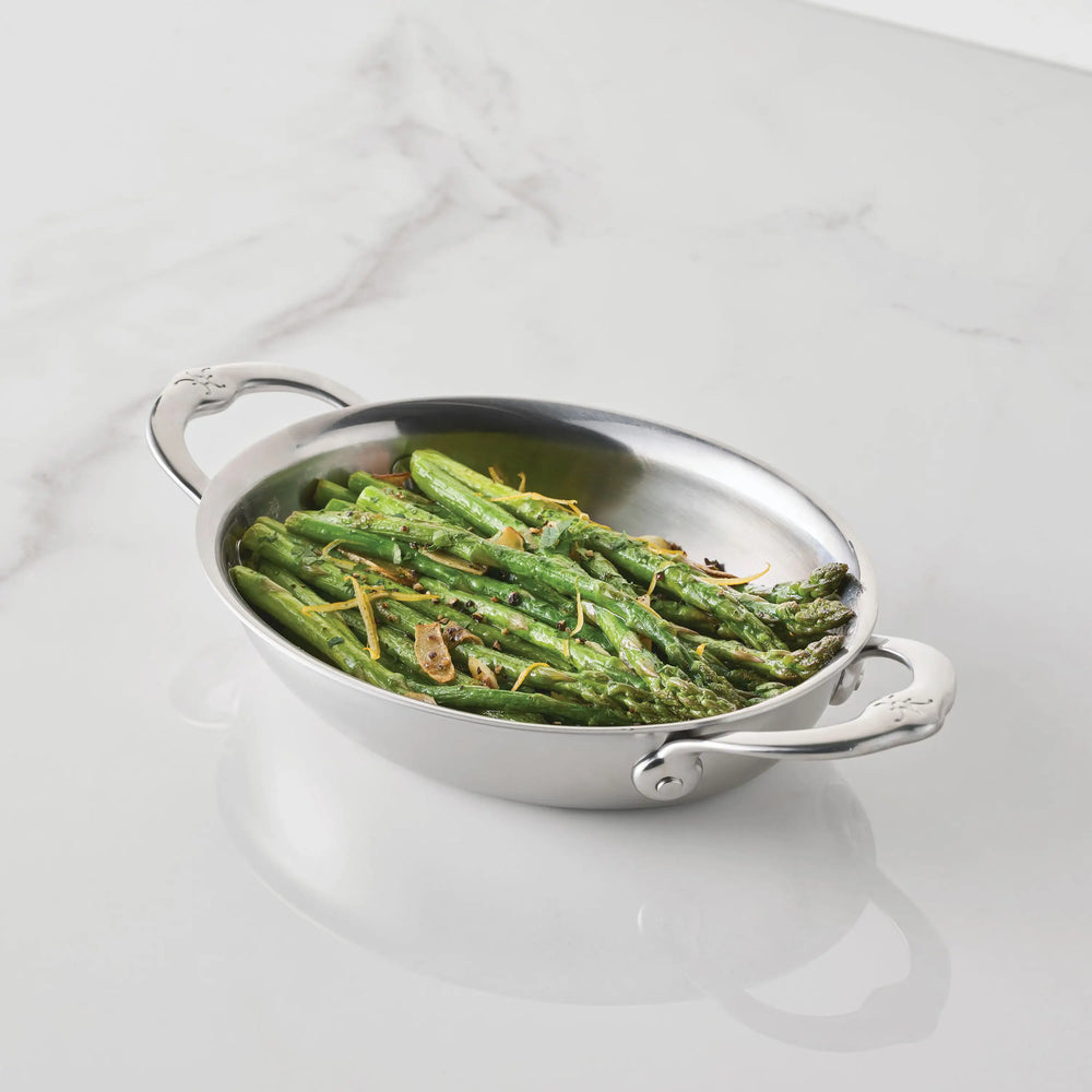 Cooked asparagus spears with sliced garlic are served in a Hestan Provisions 8.5-Inch x 6.5-Inch Oval Au Gratin dish with handles, set on a white marble surface.