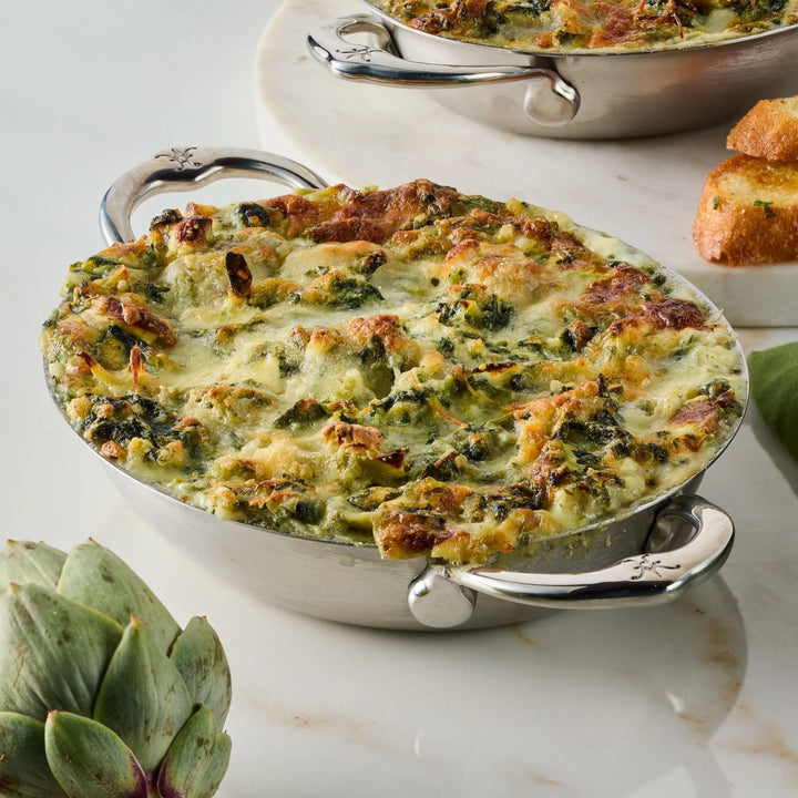 A baked spinach and artichoke dip in a Hestan Provisions 8.5-Inch x 6.5-Inch Oval Au Gratin dish sits on a marble surface, with toasted bread slices and a whole artichoke nearby.