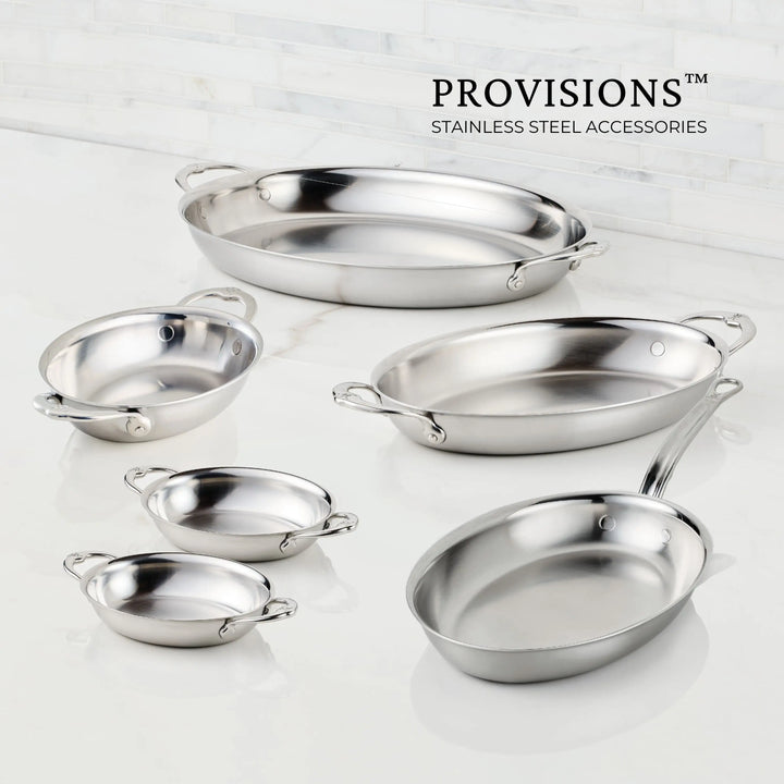 Six Hestan Provisions stainless steel pans, including the 8.5-Inch x 6.5-Inch Oval Au Gratin dish with handles, are arranged on a white countertop against a marble tile wall. The Hestan Provisions logo appears in the top right corner.