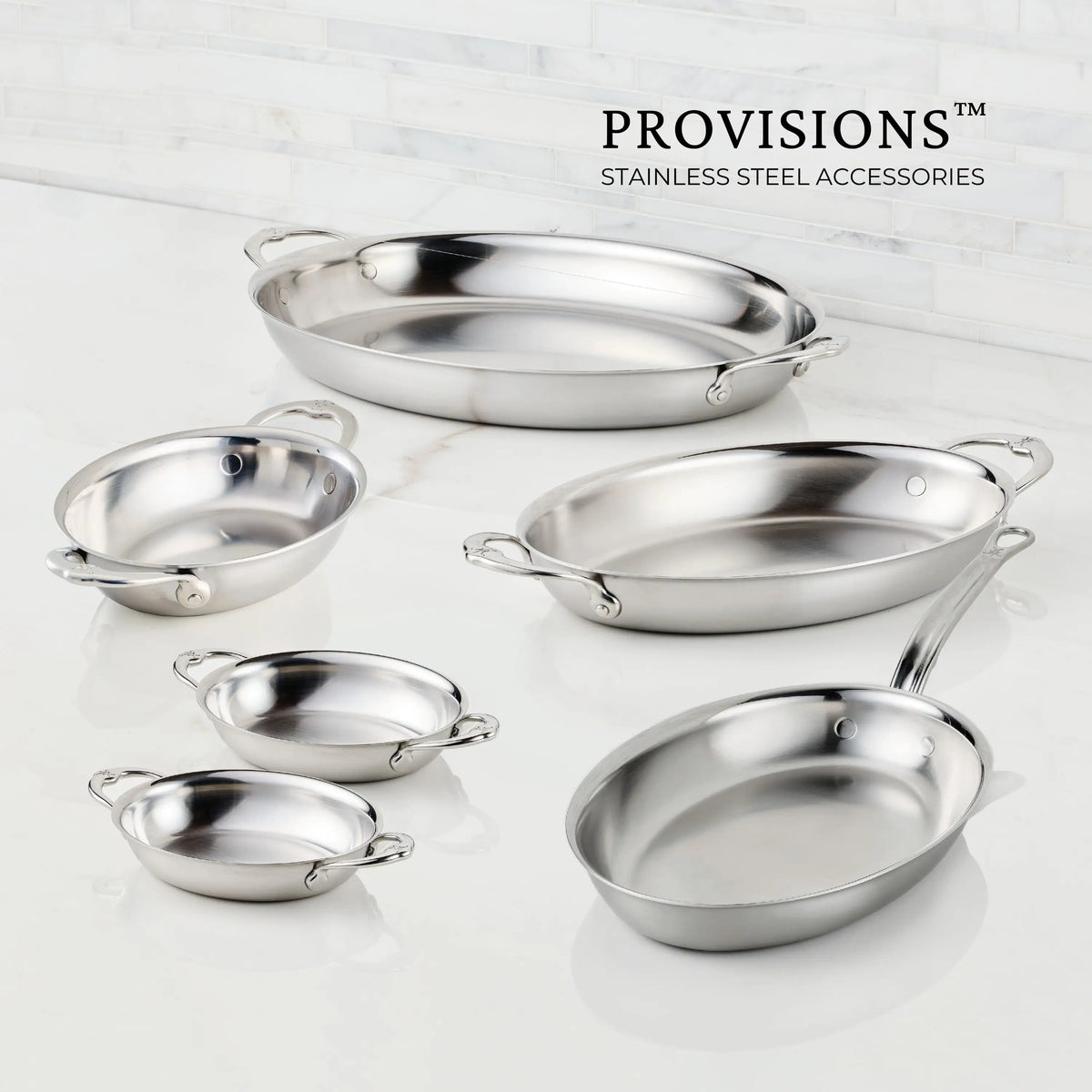 Six Hestan Provisions stainless steel pans, including the 8.5-Inch x 6.5-Inch Oval Au Gratin dish with handles, are arranged on a white countertop against a marble tile wall. The Hestan Provisions logo appears in the top right corner.