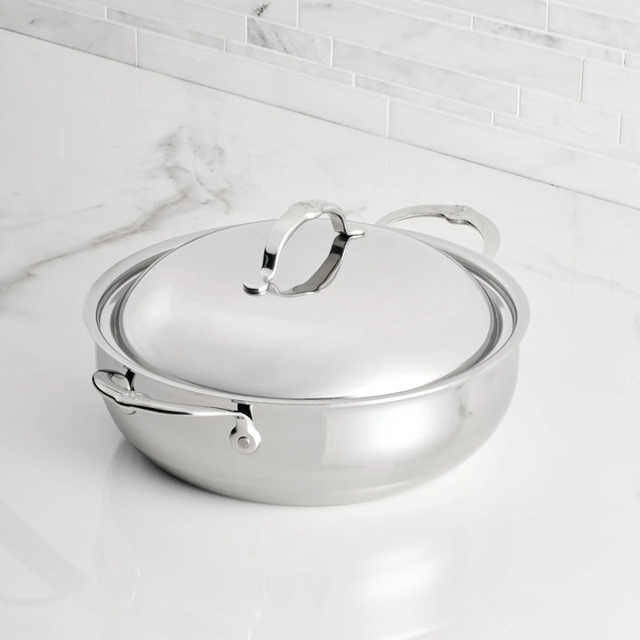 A Hestan Culinary Polished Clad Stainless Steel Dutch Oven, 5qt, with dual handles and lid, sits on a white marble countertop against a white tile backsplash.