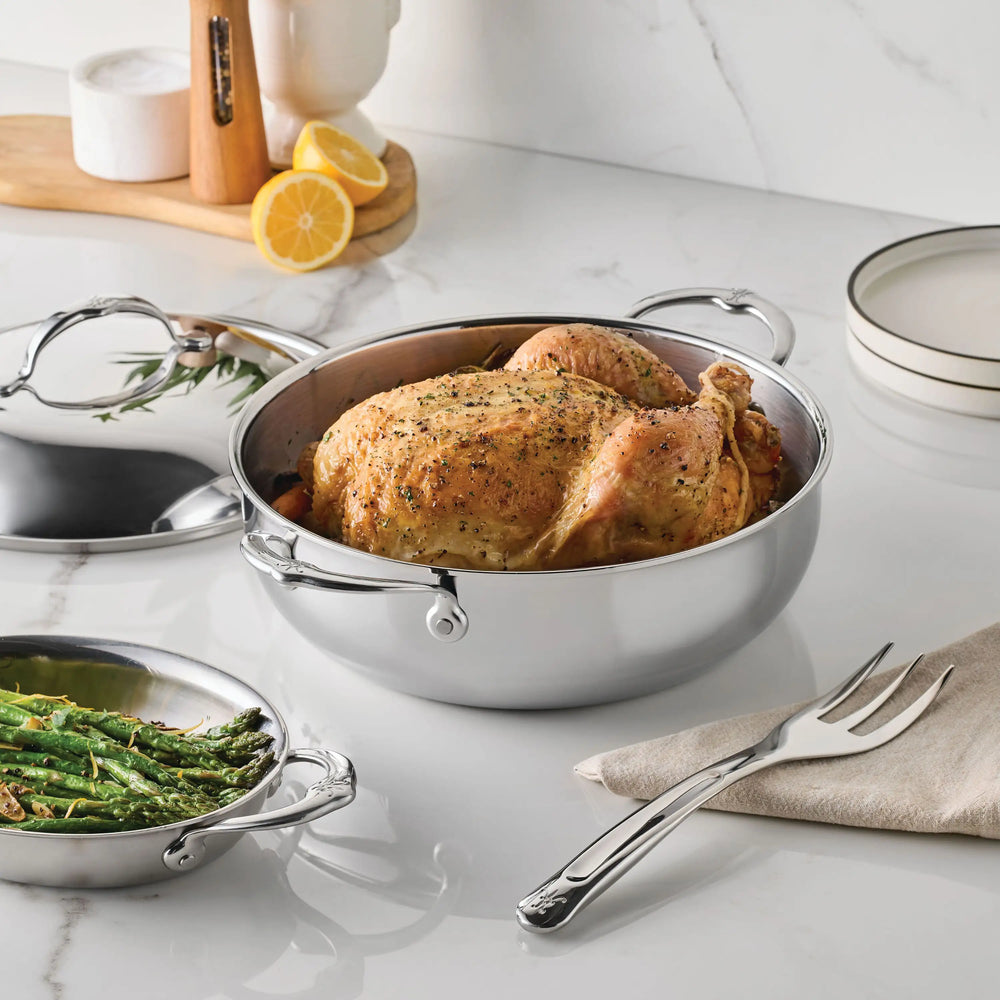A whole roasted chicken with herbs rests in a Hestan Culinary Polished Clad Stainless Steel Dutch Oven, 5qt, on a marble countertop, surrounded by a serving fork, green beans, lemon halves, plates, and utensils.