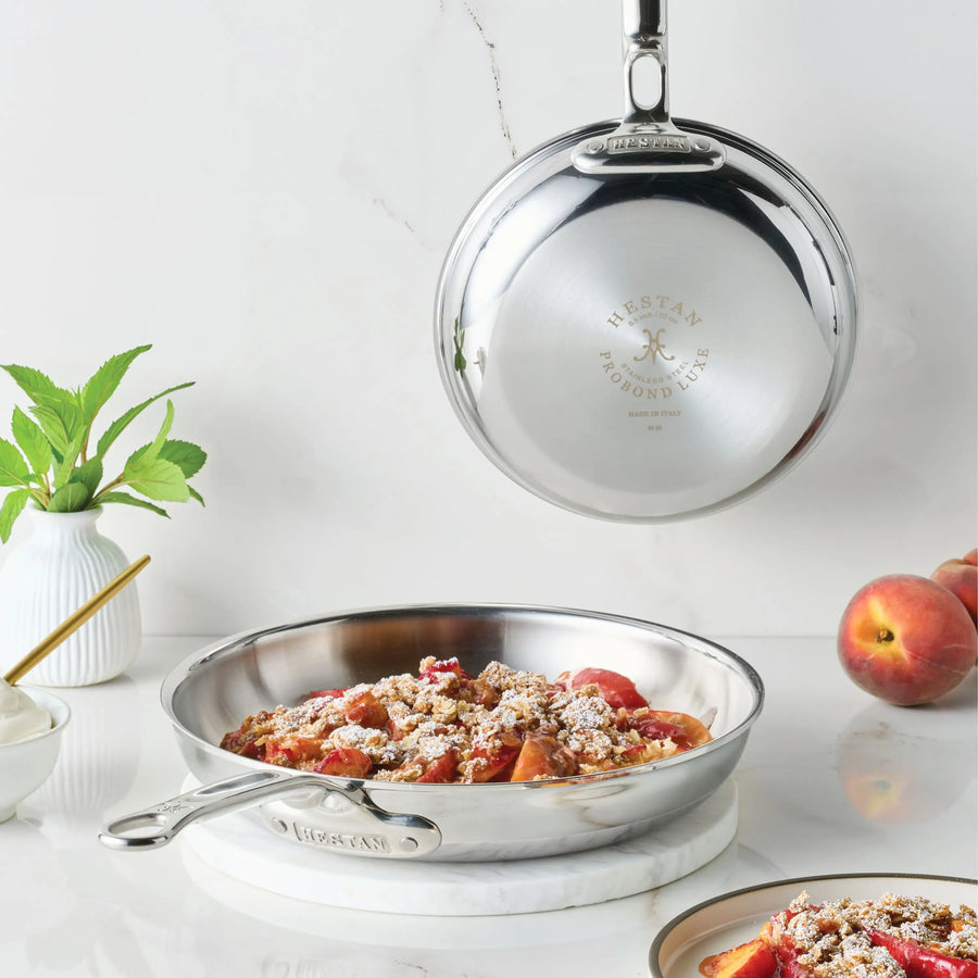 A ProBond® Luxe™ Polished Clad Stainless Steel Skillet filled with fruit dessert sits on marble, with another matching skillet hanging above it. Fresh peaches, a mint plant, and a small bowl of cream are arranged nearby.