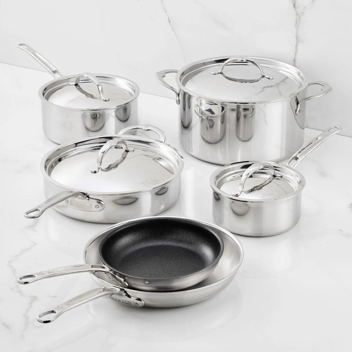 The ProBond® Luxe™ Polished Clad Stainless Steel Premiere Set, 10-Piece, showcases Italian craftsmanship with saucepans, a stockpot, sauté pan, frying pan, and nonstick skillet—each with metal handles and lids—arranged on white marble.