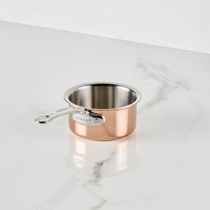 A Polished Copper 0.75-Quart Butter Warmer by CopperBond® Aura™ rests on a white marble countertop. The professional copper cookware, featuring a silver handle extending to the left, is empty and positioned slightly off-center.