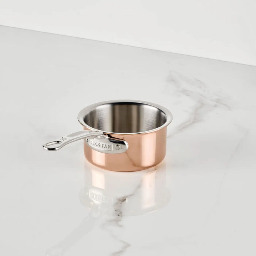 A Polished Copper 0.75-Quart Butter Warmer by CopperBond® Aura™ rests on a white marble countertop. The professional copper cookware, featuring a silver handle extending to the left, is empty and positioned slightly off-center.
