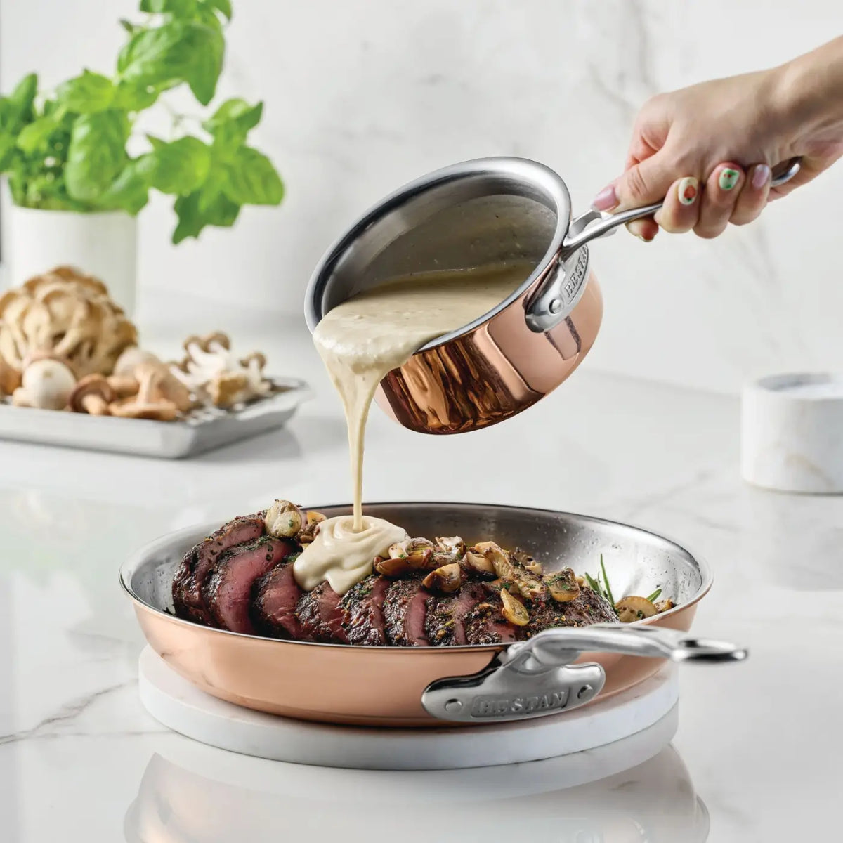 A hand pours creamy sauce from the CopperBond® Aura™ Polished Copper 0.75-Quart Butter Warmer with a stainless steel interior onto sliced steak in a copper skillet. Sautéed mushrooms top the steak, while fresh herbs and mushrooms rest on a marble countertop in the background, showcasing the use of professional copper cookware.