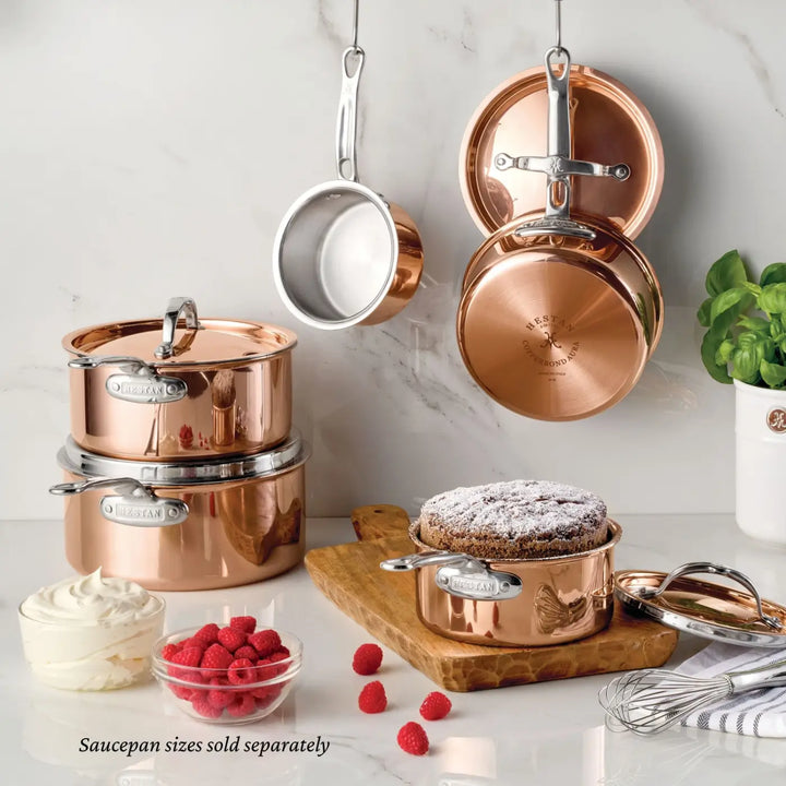 Polished CopperBond® Aura™ Saucepans with a stainless steel interior and matching lids are displayed on a counter and hanging on a wall. A cake, raspberries, whipped cream, a wooden board, and a basil plant are also present. Text at the bottom says, “Polished CopperBond® Aura™ Saucepan sizes sold separately.”.