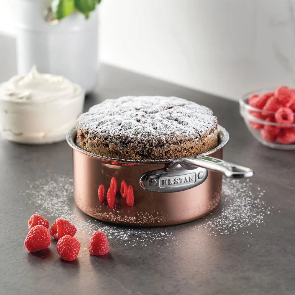 A decadent chocolate cake dusted with powdered sugar rests elegantly in a CopperBond® Aura™ Polished Copper Saucepan with Lid, highlighting the sophistication of premium copper cookware on the countertop. Fresh raspberries are scattered nearby, accompanied by a bowl of raspberries and a bowl of whipped cream in the background.