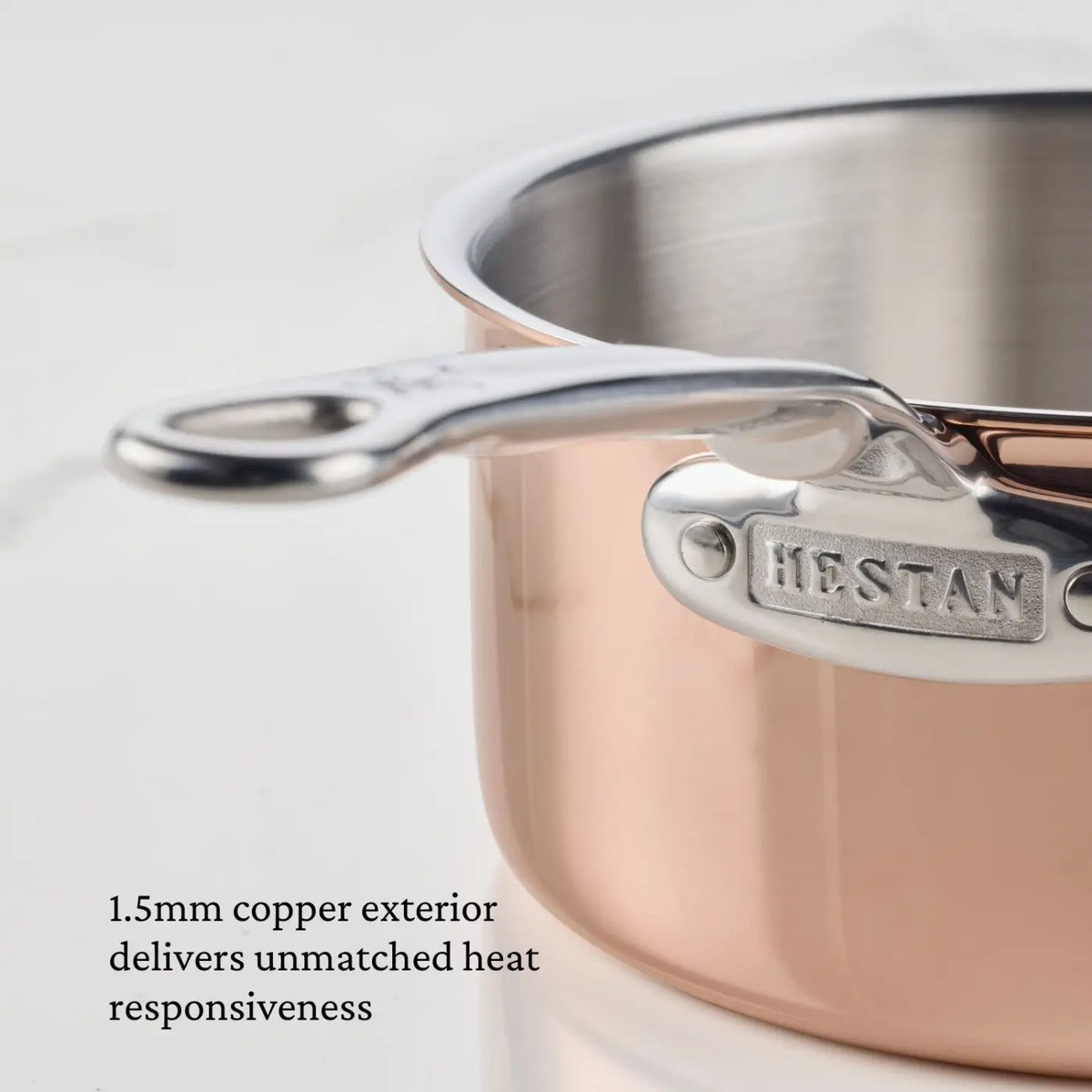 A close-up of a CopperBond® Aura™ Polished Copper Saucepan with Lid showcases its steel handle engraved with HESTAN. Text below reads: A 1.5mm copper exterior offers unrivaled heat responsiveness, while the stainless steel interior guarantees durability and easy cleanup—an elegant upgrade to your collection of premium copper cookware.