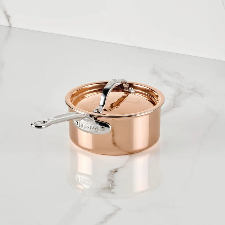 A Polished Copper Saucepan with Lid from the CopperBond® Aura™ collection sits atop a white marble surface, showcasing the refined elegance of premium copper cookware. The closed lid, engraved with the Hestan name, features a sleek stainless steel handle. Inside, its stainless steel interior promises long-lasting durability and outstanding cooking performance, set against the backdrop of a pristine white marble wall.