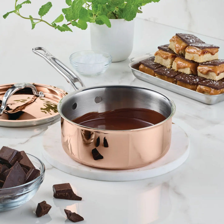 A Polished Copper Saucepan with Lid from the CopperBond® Aura™ collection, featuring a gleaming stainless steel interior, is filled with melted chocolate and sits on a white surface. Surrounding it are its matching lid, a bowl of chocolate pieces, a potted mint plant, a bowl of salt, and a tray of layered toffee bars—a stunning display of luxurious CopperBond® Aura™ copper cookware in action.