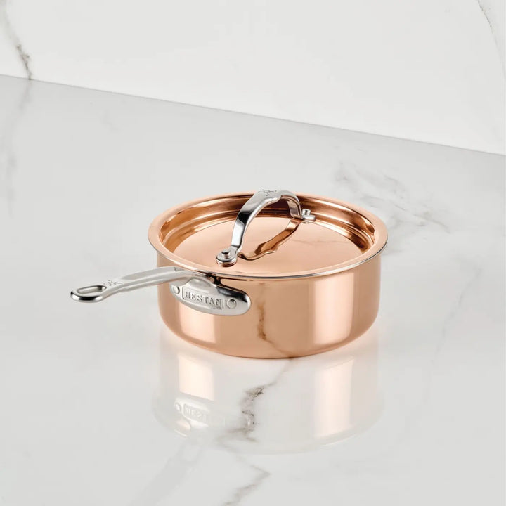 A CopperBond® Aura™ Polished Copper Saucepan with Lid, featuring a silver handle, rests on a white marble surface. This premium copper cookware item boasts a stainless steel interior, a branded metal label on the handle, and a reflective polished finish.