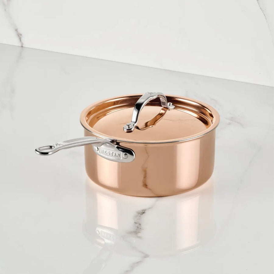 A Polished Copper Saucepan with Lid from CopperBond® Aura™ rests on a white marble countertop. This elegant piece of cookware showcases a gleaming, reflective copper surface, complemented by a silver handle and lid. Designed for durability and easy cleaning, the saucepan features a stainless steel interior, with the CopperBond® Aura™ brand name engraved on the handle. In the background, a light marble wall adds to the sophisticated setting.