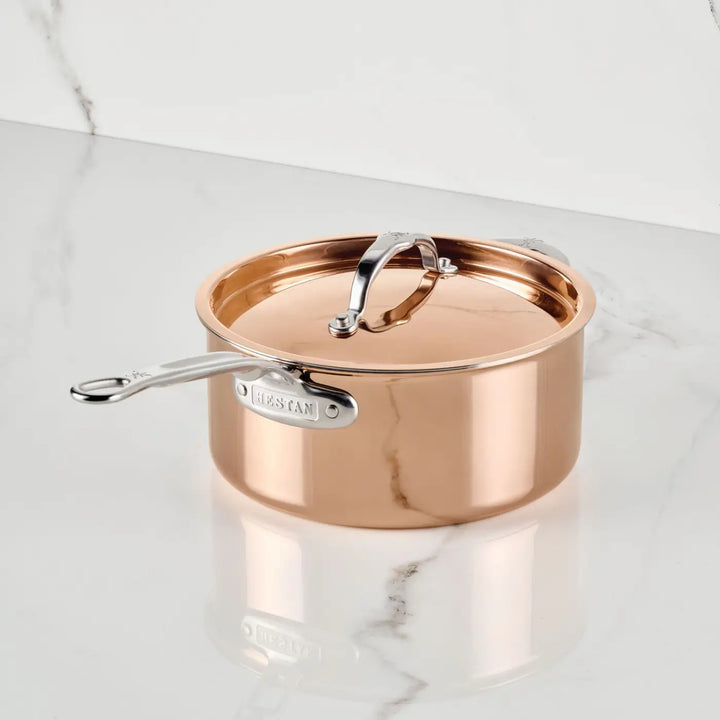 A CopperBond® Aura™ Polished Copper Saucepan with Lid sits on a white marble countertop. This premium piece features a polished copper finish, a durable stainless steel interior, a stainless steel handle and lid, and displays the CopperBond® Aura™ brand label on its side.