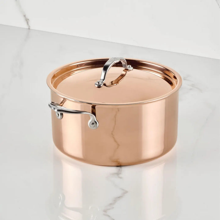 A CopperBond® Aura™ Polished Copper Stock Pot with Lid sits on a white marble surface. This striking round piece of copper cookware is highly reflective, featuring a robust stainless steel interior and sturdy stainless steel handles riveted to the sides and top of the lid.