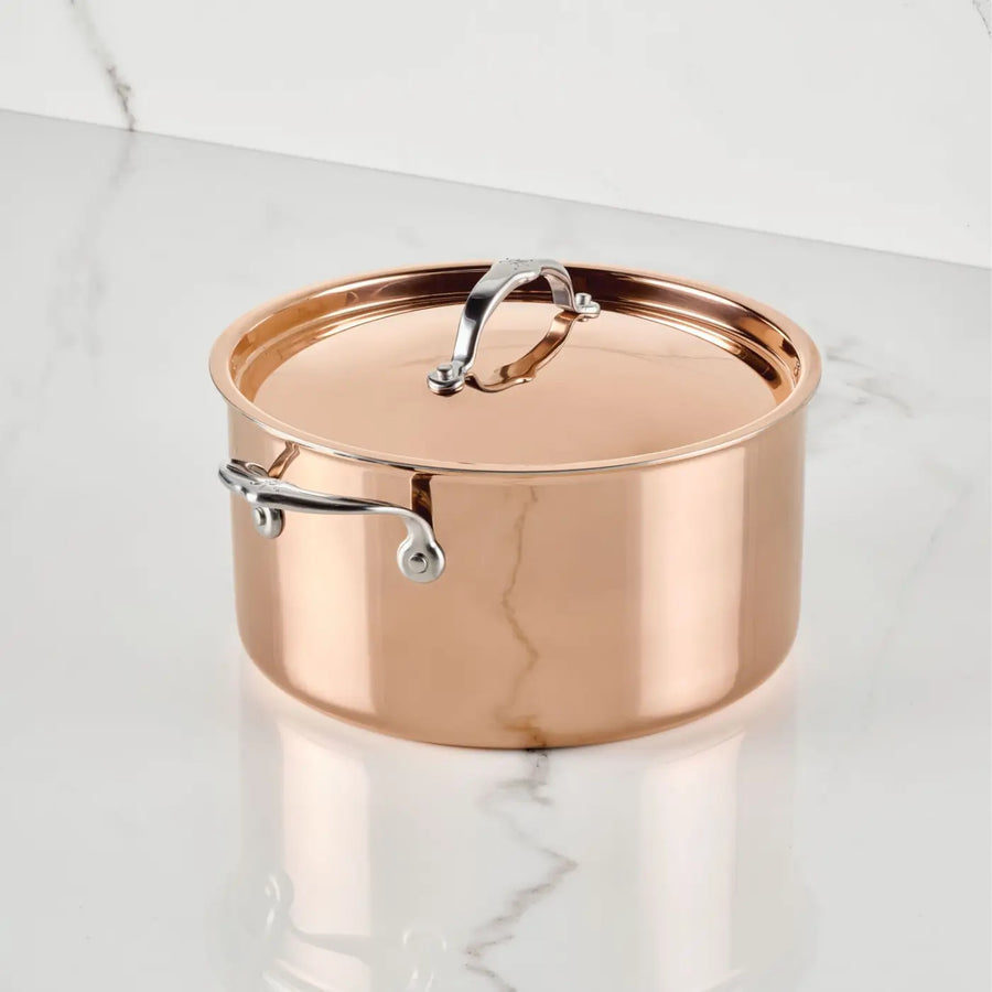A CopperBond® Aura™ Polished Copper Stock Pot with Lid sits on a white marble surface. This striking round piece of copper cookware is highly reflective, featuring a robust stainless steel interior and sturdy stainless steel handles riveted to the sides and top of the lid.