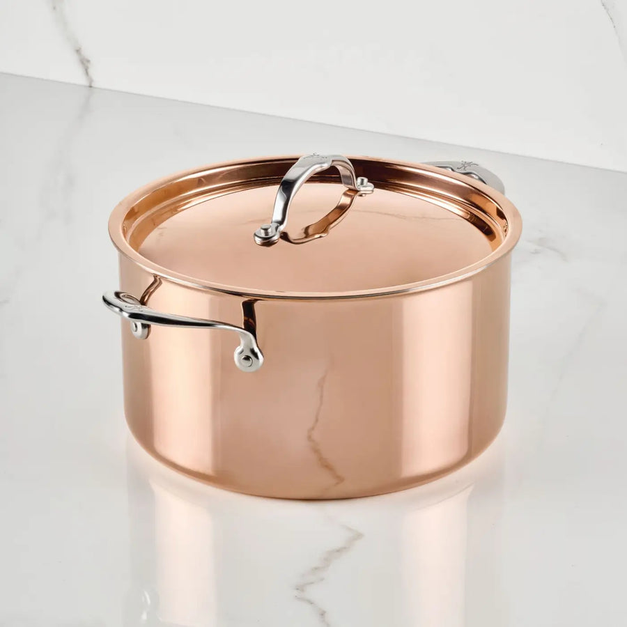 A Polished Copper Stock Pot with Lid from CopperBond® Aura™ sits on a white marble surface with gray veining. This elegant cookware showcases its lustrous copper finish and sleek stainless steel handles, reflecting light to highlight its sophisticated design.