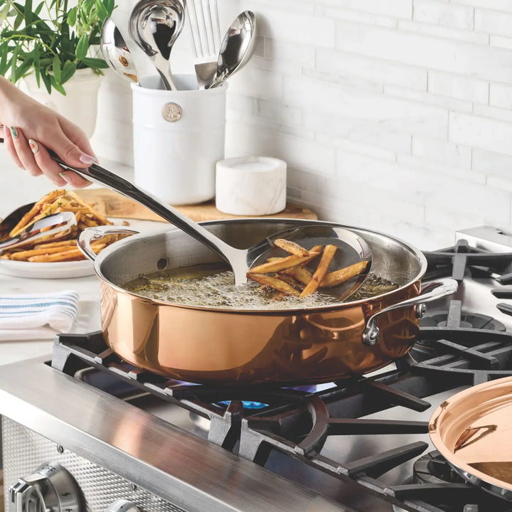 A hand uses a slotted spoon to lift French fries from hot oil in the CopperBond® Aura™ Polished Copper Rondeaus with Lid on a gas stove, taking advantage of its excellent heat responsiveness. Cooked fries rest on a plate nearby, while stainless steel utensils and a plant are visible in the background.