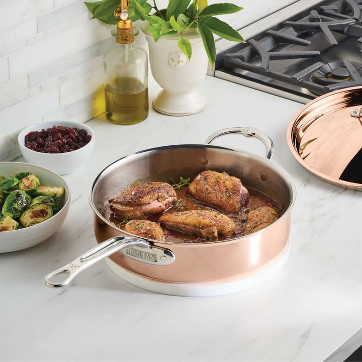 A Polished Copper Saute Pan with Lid from the CopperBond® Aura™ collection—featuring a sleek stainless steel interior and filled with cooked, seasoned chicken pieces—sits on a marble counter beside a bowl of Brussels sprouts, a small bowl of cranberries, an olive oil bottle, and a plant near a gas stove.
