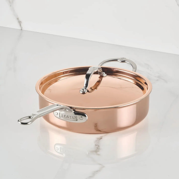 A Polished Copper Saute Pan with Lid from the CopperBond® Aura™ collection sits on a white marble surface. This pan, inspired by Italian copper cookware tradition, boasts a shiny finish, durable stainless steel interior, and features the CopperBond® Aura™ brand name on its handle.