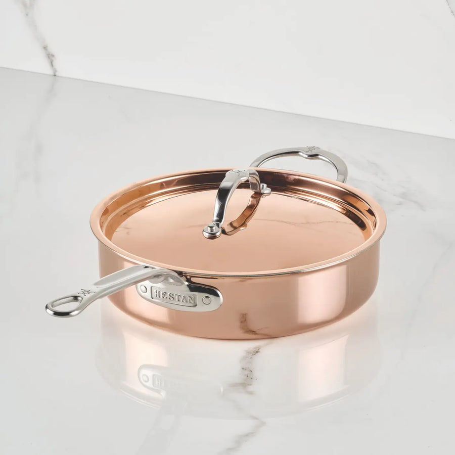 A Polished Copper Saute Pan with Lid from the CopperBond® Aura™ collection sits on a white marble surface. This pan, inspired by Italian copper cookware tradition, boasts a shiny finish, durable stainless steel interior, and features the CopperBond® Aura™ brand name on its handle.