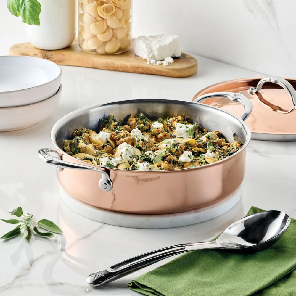 A Polished Copper Rondeau with Lid from CopperBond® Aura™, renowned for its heat responsiveness, sits on a marble countertop near a green napkin and spoon. The stainless steel interior is filled with cooked pasta, vegetables, and chunks of cheese, while a jar of pasta, a block of cheese, and a bowl appear in the background.