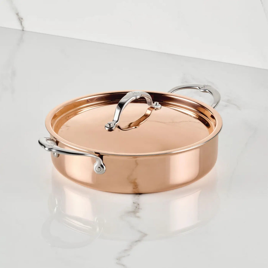 A CopperBond® Aura™ Polished Copper Rondeaus with Lid rests on a white marble surface with gray veining. Esteemed for its exceptional heat responsiveness, this copper cookware piece boasts a durable stainless steel interior and sturdy stainless steel handles. The securely placed lid and the polished pan reflect surrounding light, highlighting its elegant design.