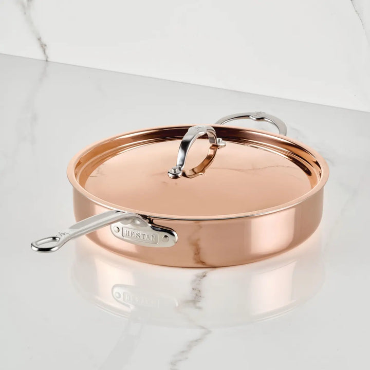 A Polished Copper Saute Pan with Lid from CopperBond® Aura™, featuring a gleaming stainless steel interior and two silver handles, rests on a white marble countertop. The brand name is elegantly displayed on the side handle, reflecting the refined craftsmanship of Italian-inspired copper cookware.