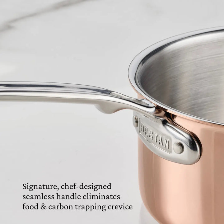 Close-up of a professional-grade Polished Copper Essential Pan with Lid from CopperBond® Aura™, featuring a seamless stainless steel handle securely attached by rivets and an embossed Made In mark. Text reads: Signature, chef-designed seamless handle eliminates food & carbon trapping crevice—an ideal example of Italian crafted cookware excellence.