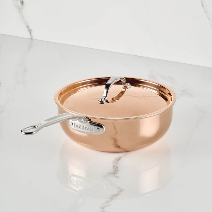 The CopperBond® Aura™ Polished Copper Essential Pan with Lid, featuring a silver handle engraved with the brand name and a coordinating copper lid, rests elegantly on a white marble surface adorned with grey veining—an exquisite example of Italian-crafted cookware.