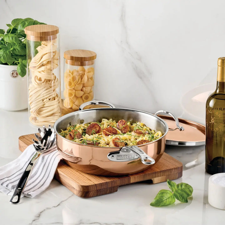 A CopperBond® Aura™ Polished Copper Essential Pan with Lid, filled with pasta and vegetables, rests on a wooden board. Surrounding it are a striped towel, a pasta server, jars of dry pasta, a bottle of white wine, a plant, and basil leaves on the marble countertop—a perfect setting to showcase this Italian-crafted cookware that brings authentic elegance to your kitchen.