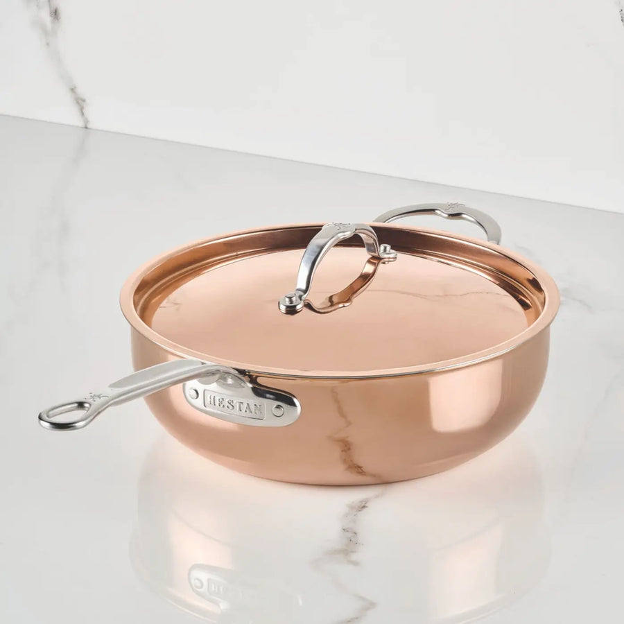 A Polished Copper Essential Pan with Lid from CopperBond® Aura™ sits on a white marble countertop. The pan’s reflective surface and sturdy metal handles highlight the elegance of professional-grade copper cookware in your kitchen.