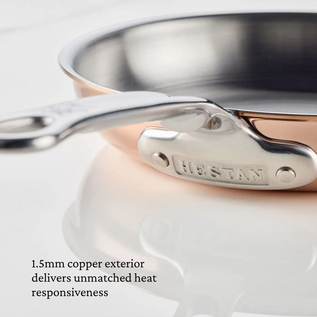 Here’s a rewritten version using your specified product data: Close-up of a CopperBond® Aura™ Polished Copper Skillet handle. Text on image reads: 1.5mm copper exterior delivers unmatched heat responsiveness. This professional-grade skillet features a stainless steel interior and rests on a reflective white surface.